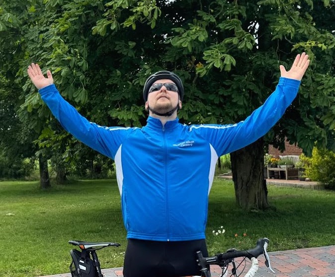 Radfahrer in blauem Trikot mit ausgebreiteten Armen neben seinem Rennrad im Grünen